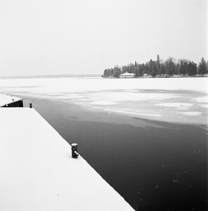 A snow-covered dock extends into a partially frozen lake with a house and trees on the far shore.