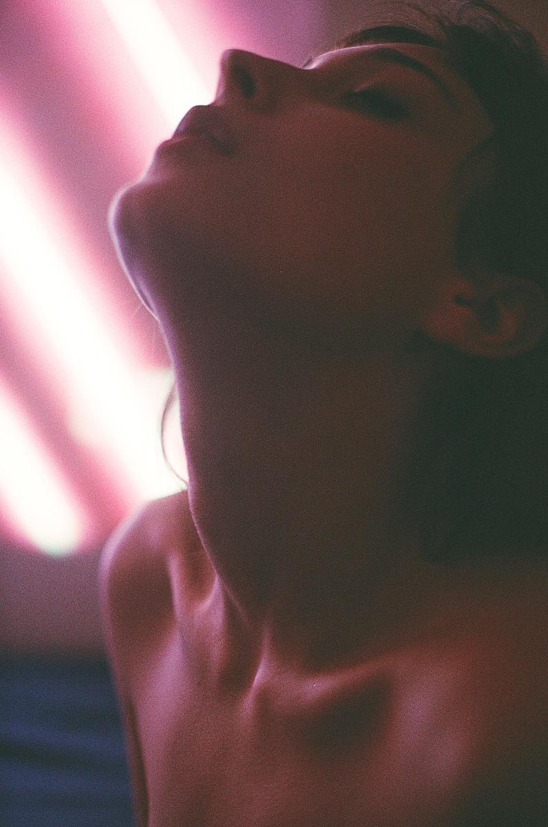 A woman's head is tilted back, with her face and neck illuminated by pink light.