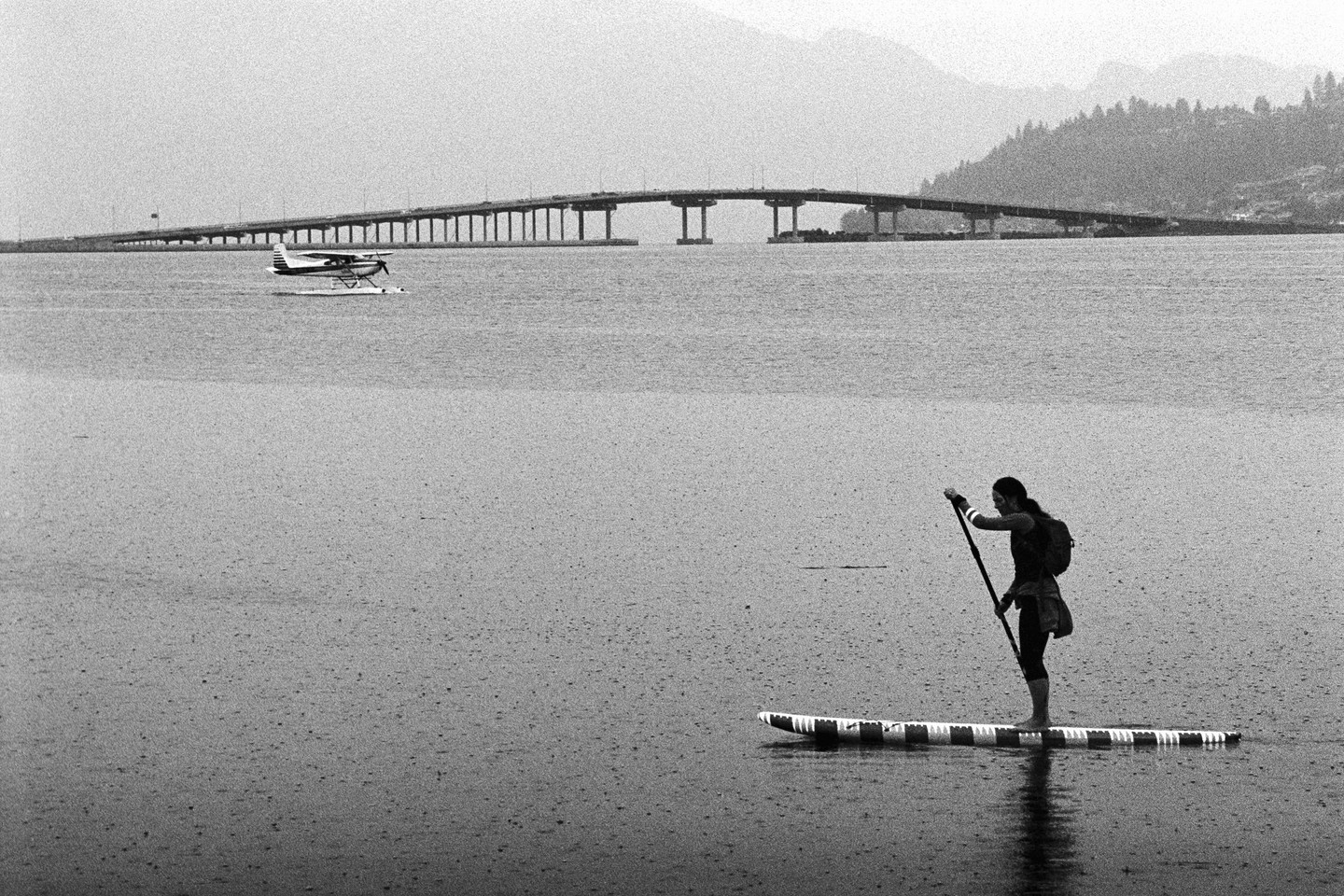 A woman paddleboards in the rain with a floatplane and a bridge in the background.