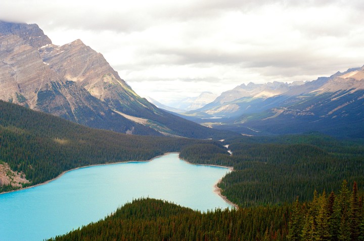A turquoise lake is surrounded by a dense forest and mountains under a cloudy sky.