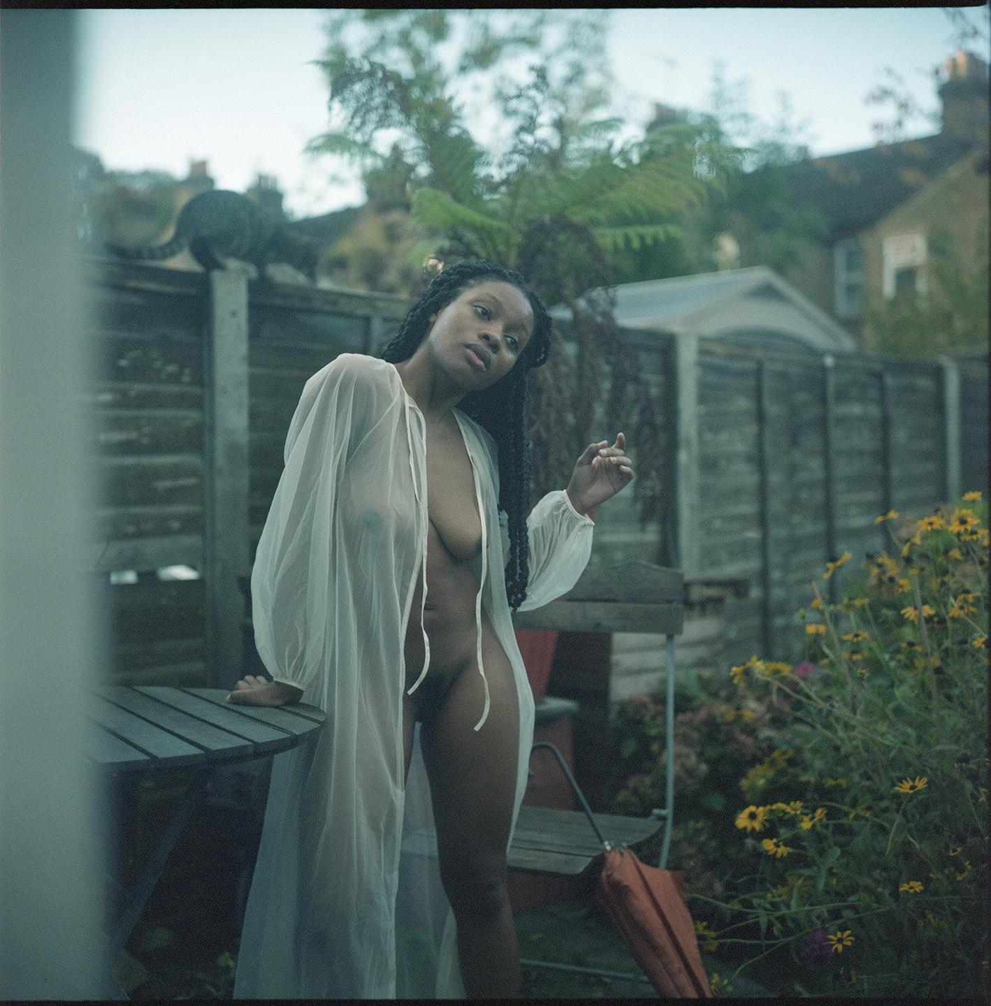 A woman in a sheer robe stands next to a table in a garden with a cat on a fence in the background.