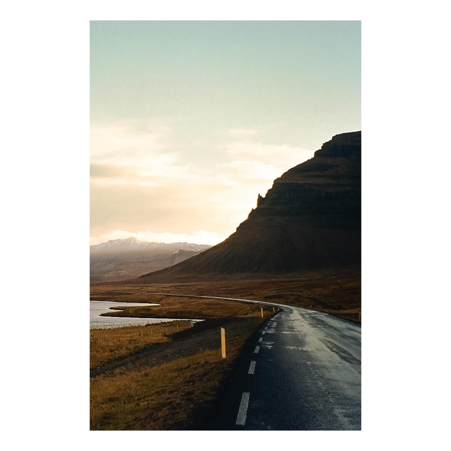 A winding road leads through a valley with a mountain on the right and a body of water on the left under a cloudy sky.