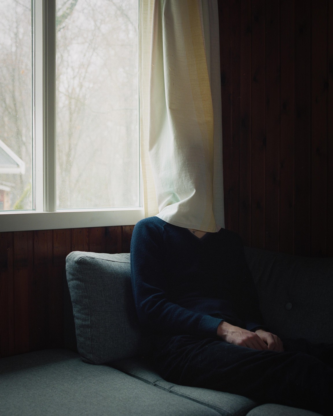 A person sits on a sofa with a curtain covering their head, next to a window looking out at bare trees.