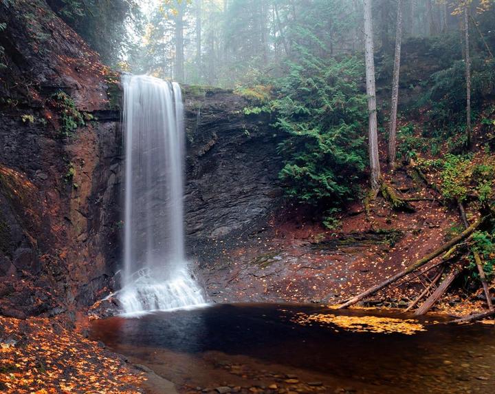 A waterfall cascades into a dark pool surrounded by autumn foliage and trees.