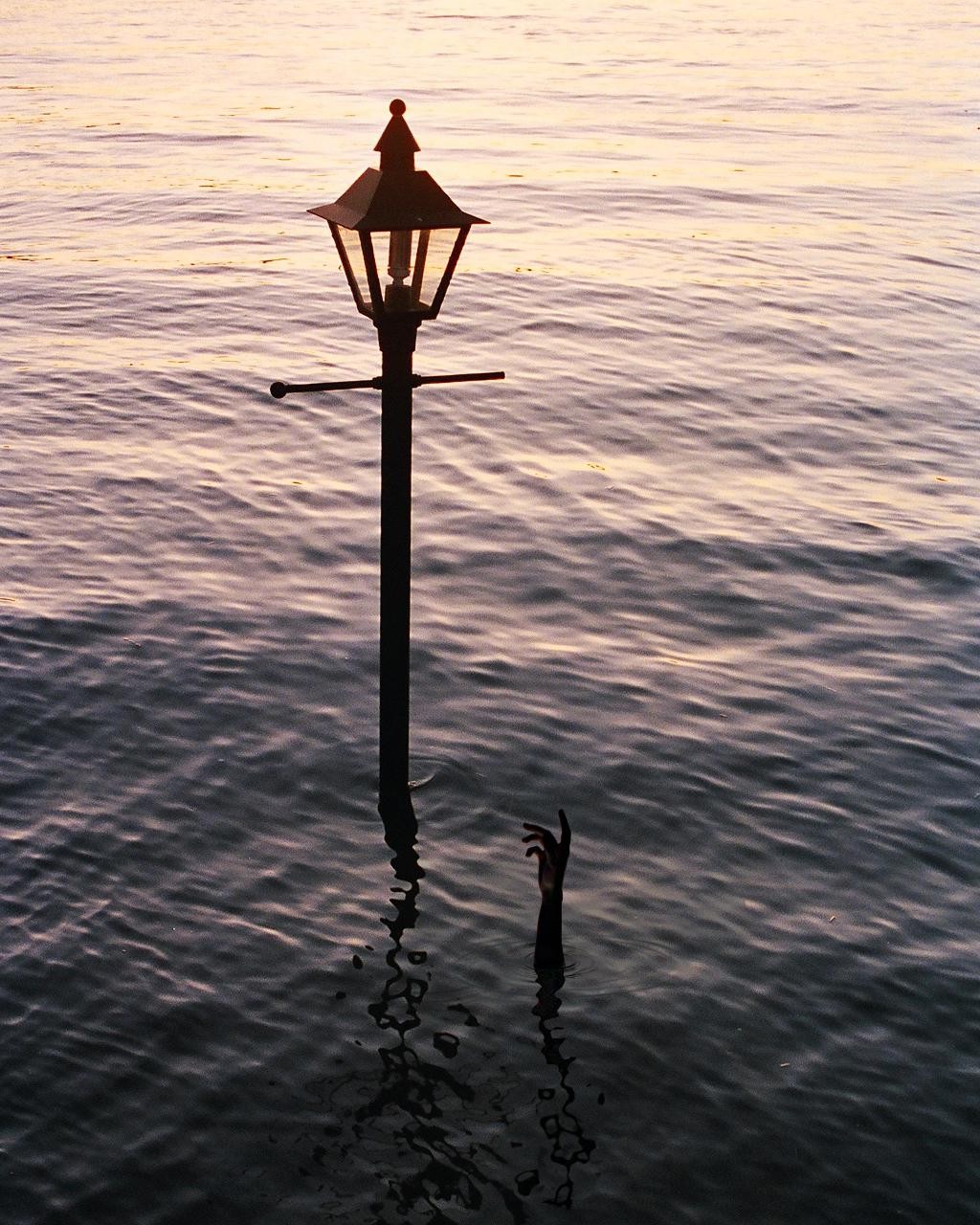 A street lamp stands in water with a hand reaching out of the water next to it.