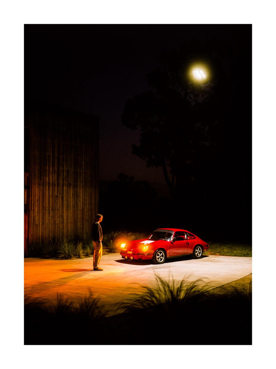 A man stands next to a red car with its headlights on at night.