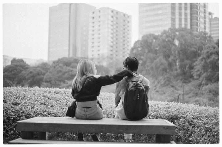A woman and a man sit on a bench with their backs to the camera, with the woman's arm around the man's shoulder, in a park with buildings in the background.