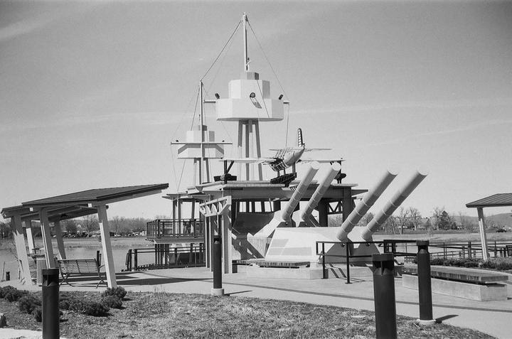A black and white photo shows a memorial park with a battleship-like structure and a small airplane.