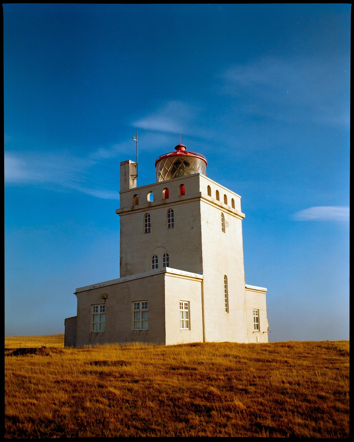 A white lighthouse with a red top stands on a hill of dry grass under a clear blue sky.