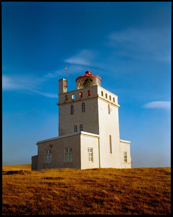 A white lighthouse with a red top stands on a hill of dry grass under a clear blue sky.