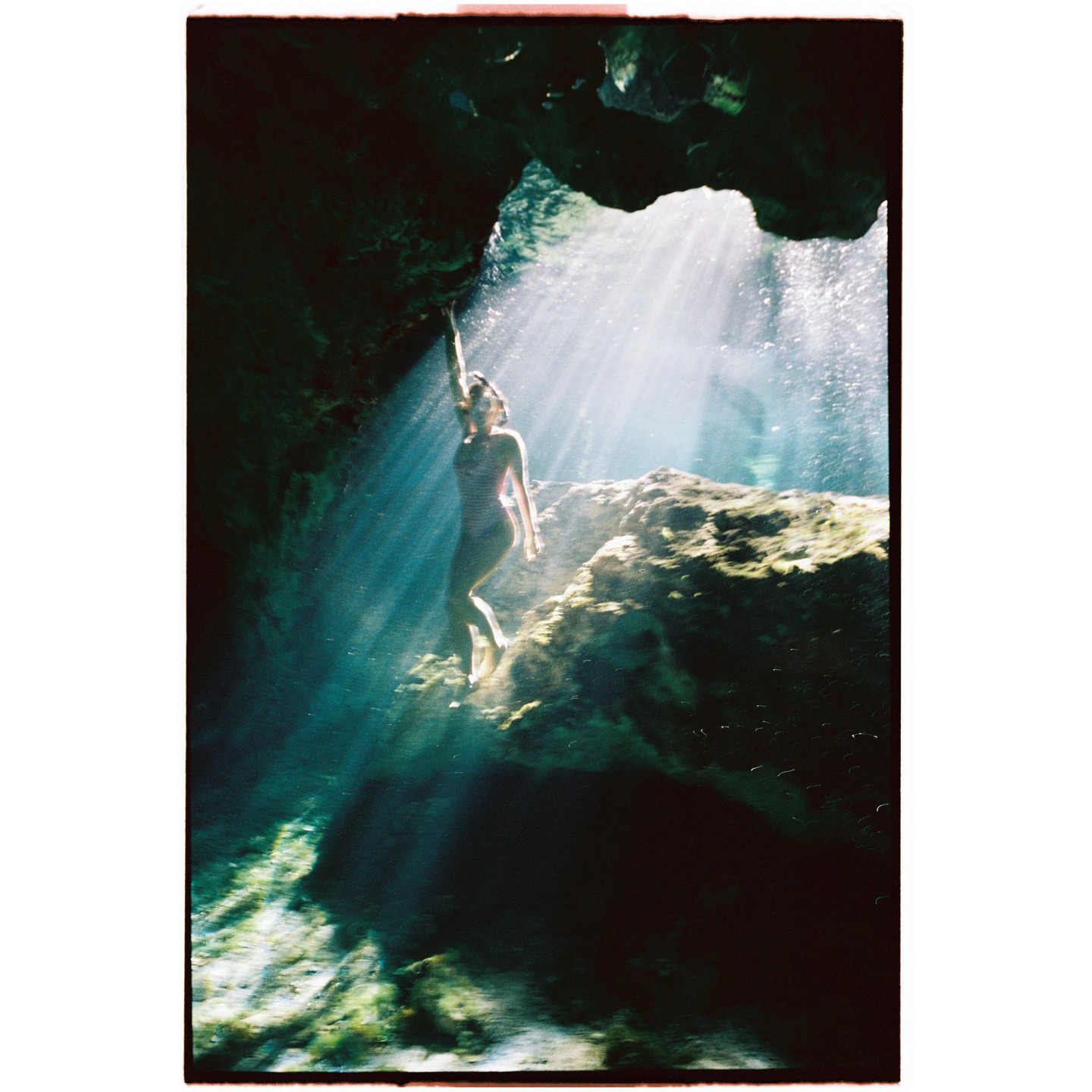 A woman in a swimsuit stands on a rock in a cave with light rays shining down on her.