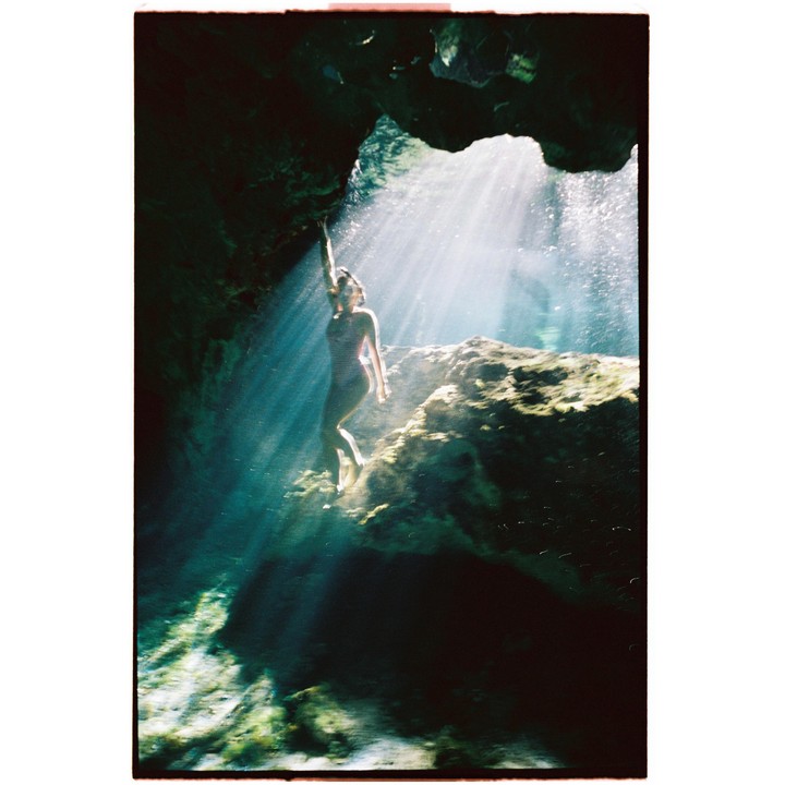 A woman in a swimsuit stands on a rock in a cave with light rays shining down on her.