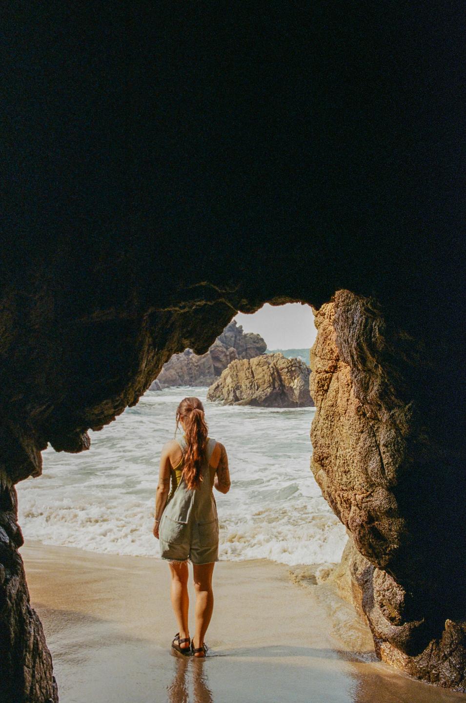 A woman stands on a sandy beach looking out at the ocean through a cave opening.