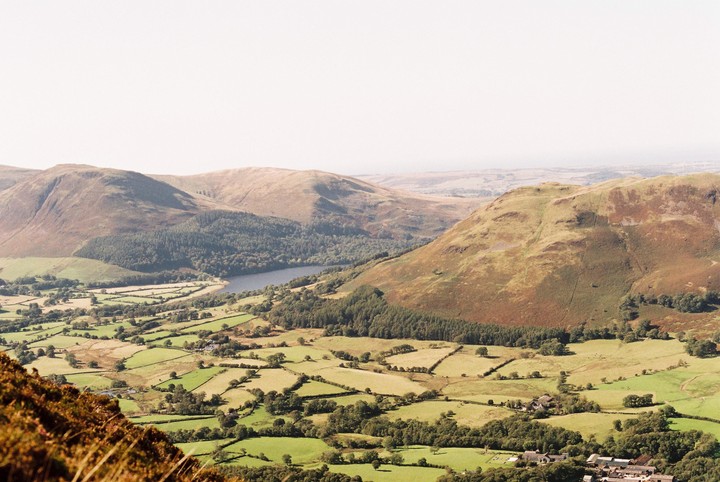 A vast landscape shows a valley with a lake, fields, and mountains under a clear sky.
