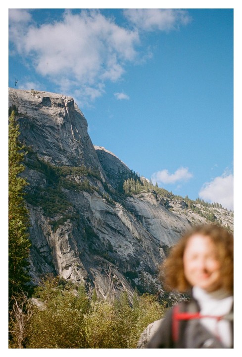 A blurry person with curly hair is in the foreground of a mountain landscape with a clear blue sky.