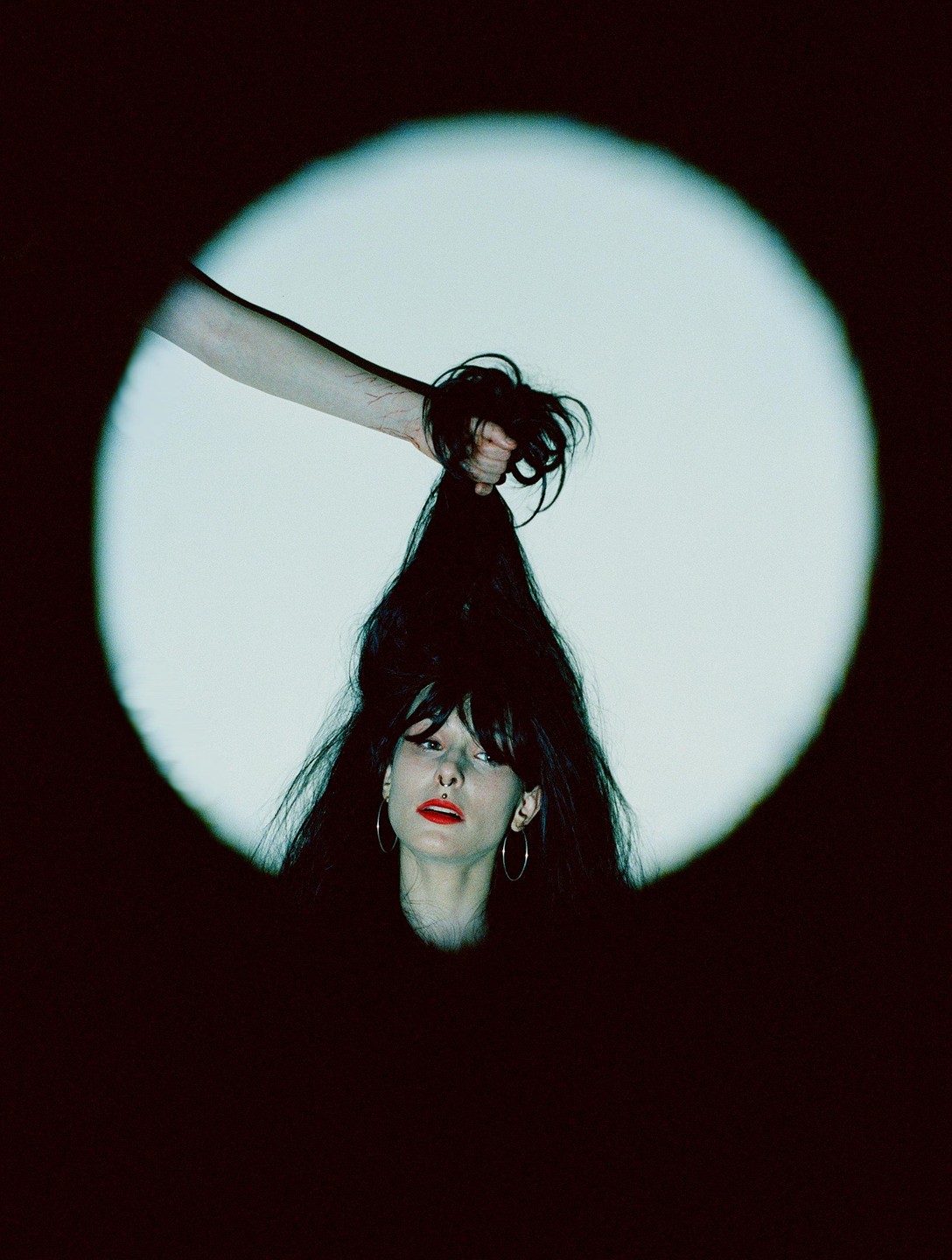 A woman with long dark hair and red lipstick is illuminated by a spotlight while a hand pulls her hair.