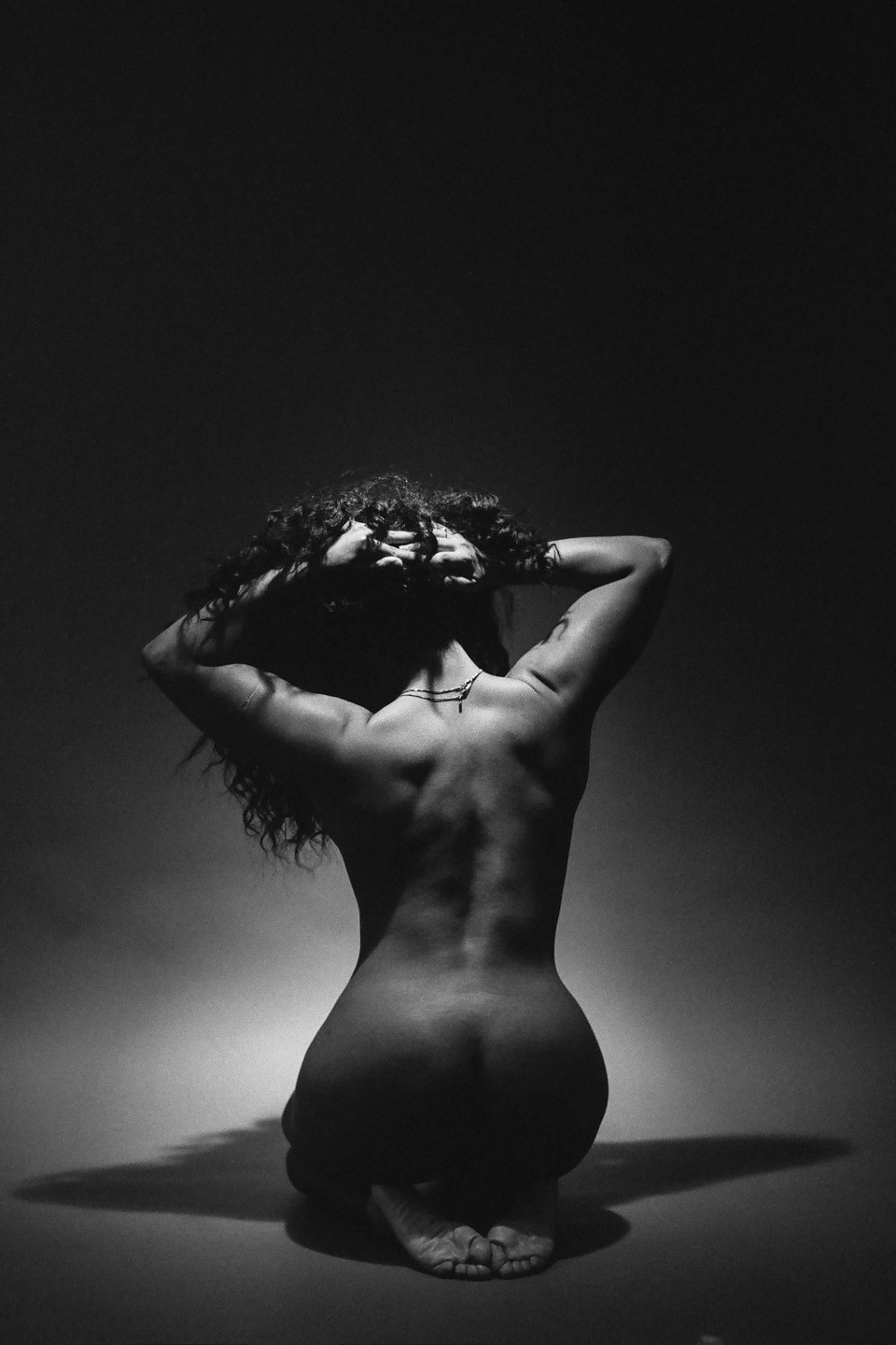 A woman with curly hair kneels with her back to the viewer, her hands behind her head.