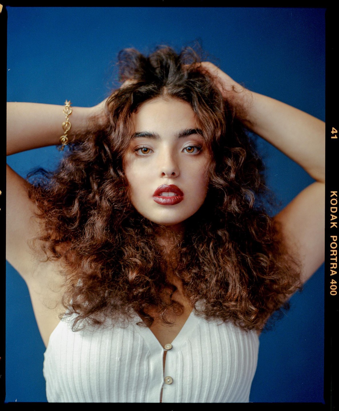 A woman with curly hair and dark red lipstick poses against a blue background, holding her hands in her hair.