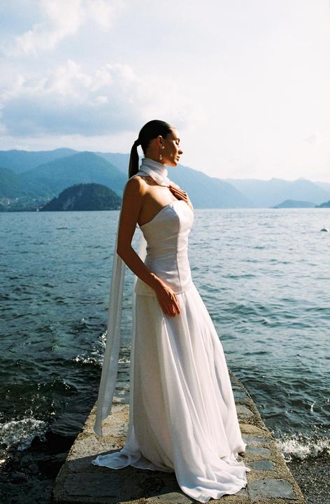 A woman in a white dress stands on a stone pier by a lake with mountains in the background.