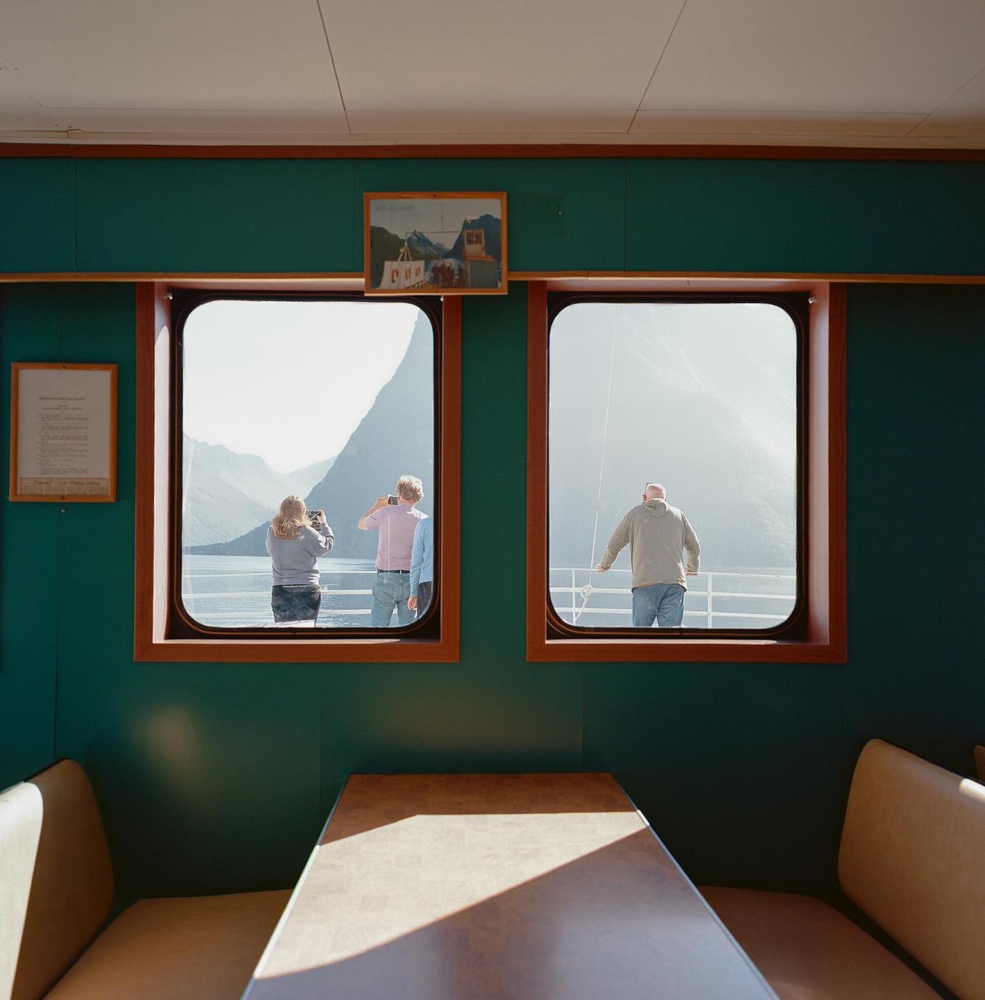 Three people are visible through two windows of a ferry, with mountains and water in the background.