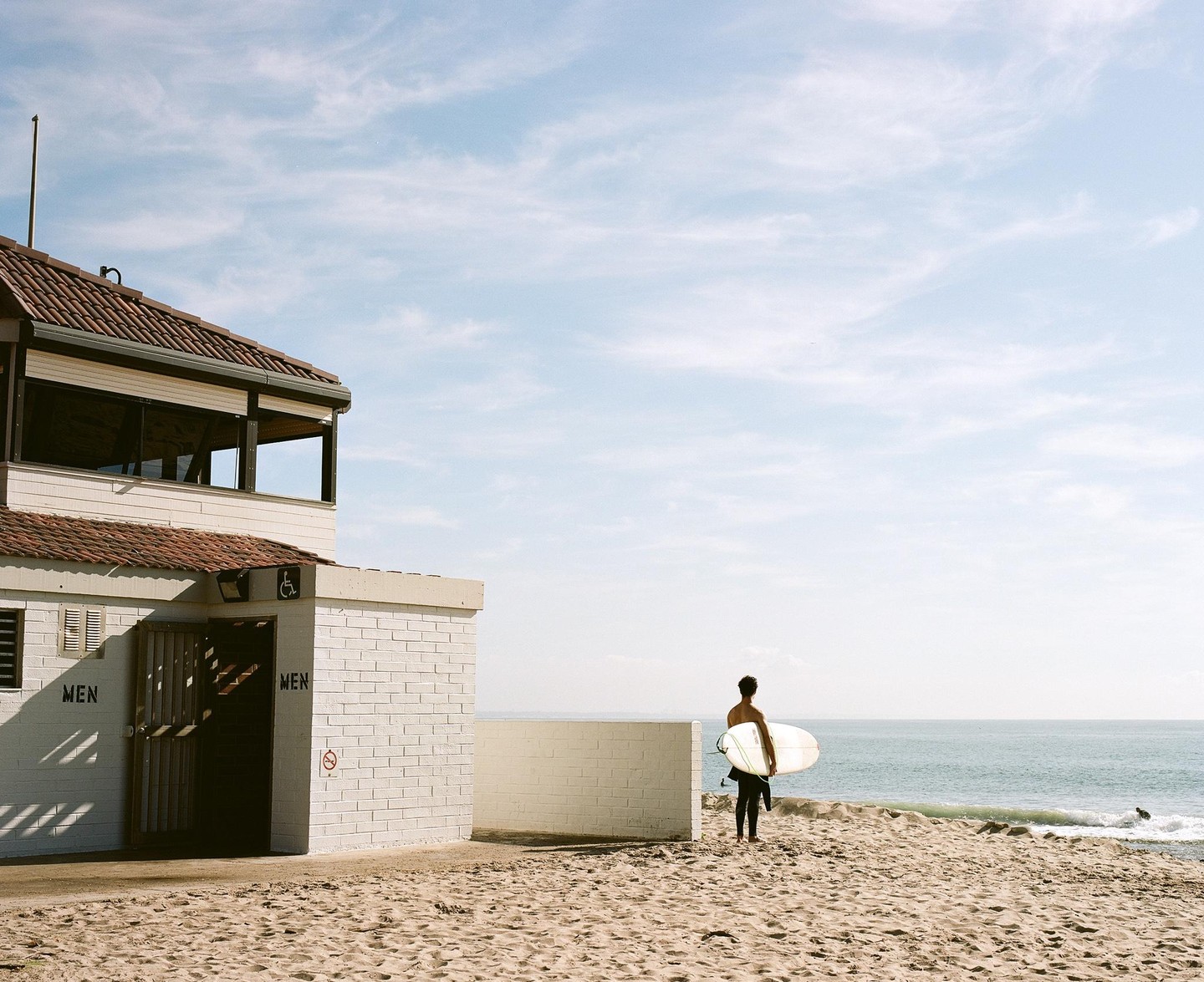 A surfer stands on a sandy beach next to a building, looking out at the ocean.