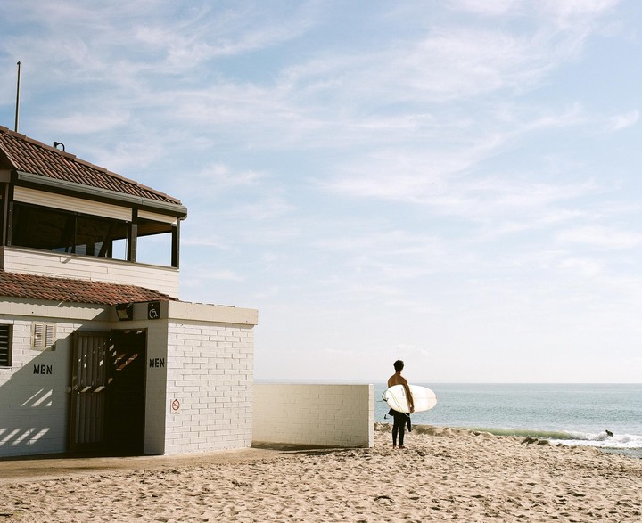A surfer stands on a sandy beach next to a building, looking out at the ocean.