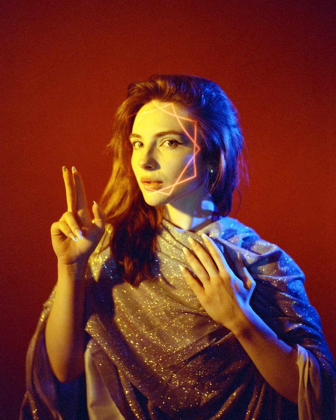 A woman with a glowing geometric pattern on her face holds up two fingers and wears a shimmering garment.