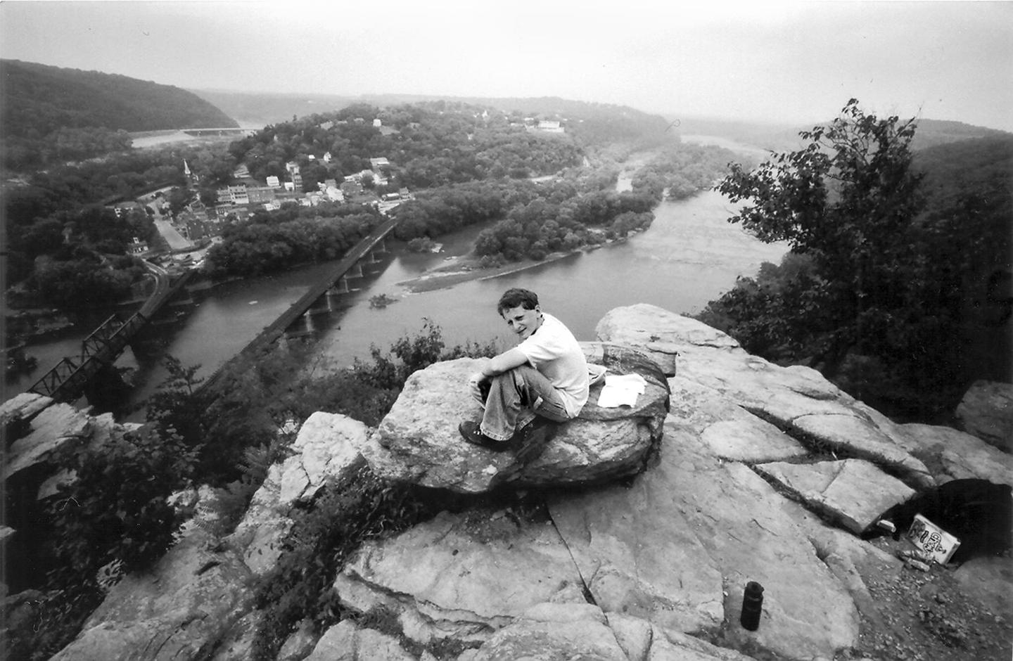 A man sits on a rock overlooking a river with bridges and a town nestled among trees.