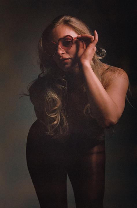 A woman with long blonde hair is wearing heart-shaped sunglasses and looking towards the viewer.