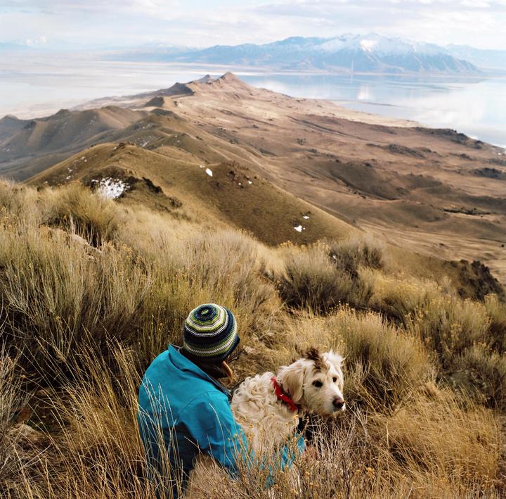 A person and a dog sit in tall grass on a hillside overlooking a lake and mountains.