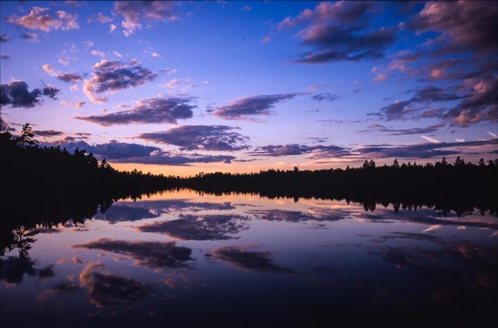 Clouds are reflected in a lake at sunset, with a dark forest silhouetted on the far bank.