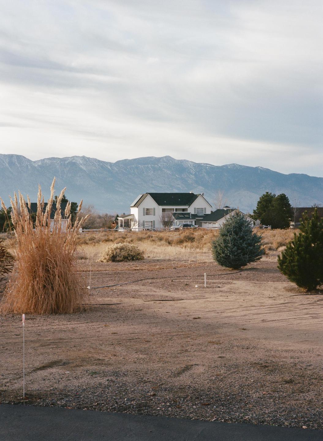 A white house with a dark roof stands in a dry, grassy field with mountains in the background.