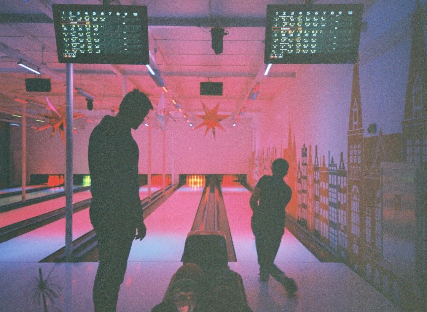 Three people are bowling in a pink-lit alley with a city mural on the wall.