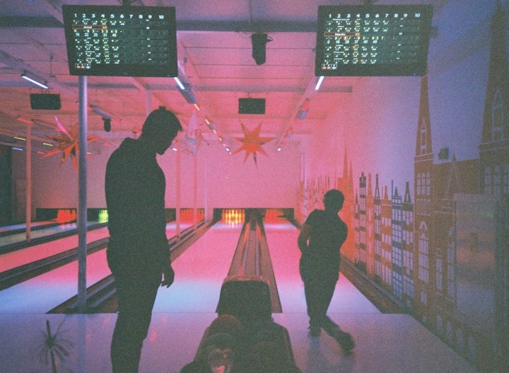 Three people are bowling in a pink-lit alley with a city mural on the wall.