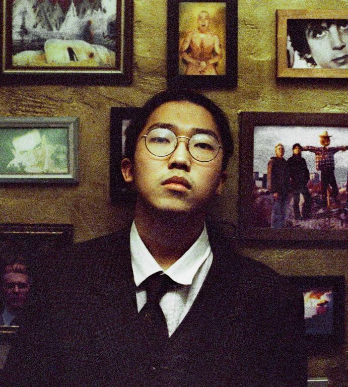A young man wearing glasses, a white shirt, and a dark tie and jacket stands in front of a wall covered with framed pictures.