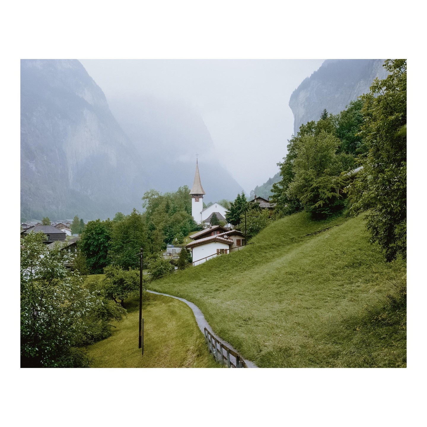 A small village with a church and houses is nestled in a valley surrounded by mountains and green hills.