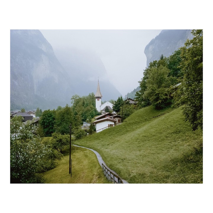 A small village with a church and houses is nestled in a valley surrounded by mountains and green hills.