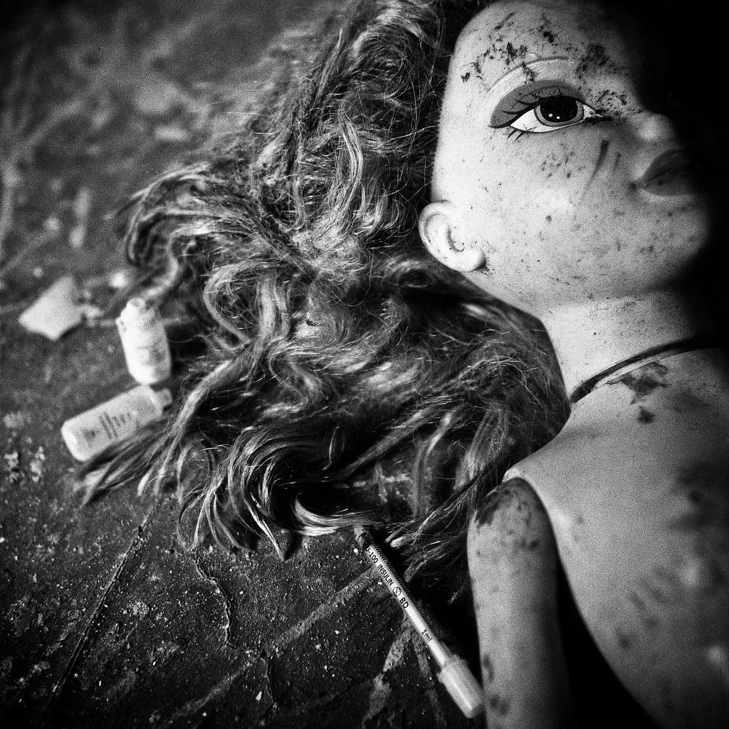 A dirty doll head with long, curly hair lies on a grimy surface next to a syringe and small bottles.