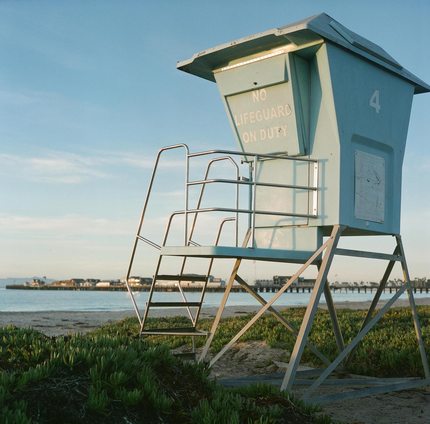 A light blue lifeguard stand with a sign that reads "No Lifeguard On Duty" stands on a sandy beach with green plants in the foreground and a pier in the background.