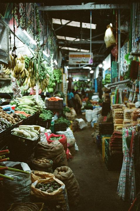 A narrow market stall is filled with produce and sacks of goods under a corrugated roof.