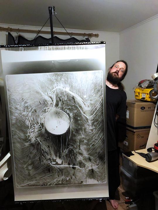 A man with a beard peeks from behind a large, abstract print hanging in a room.