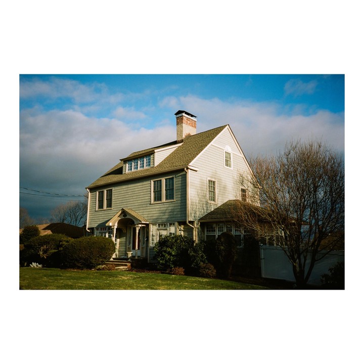 A large house with light blue siding and a brown roof stands on a green lawn under a partly cloudy sky.
