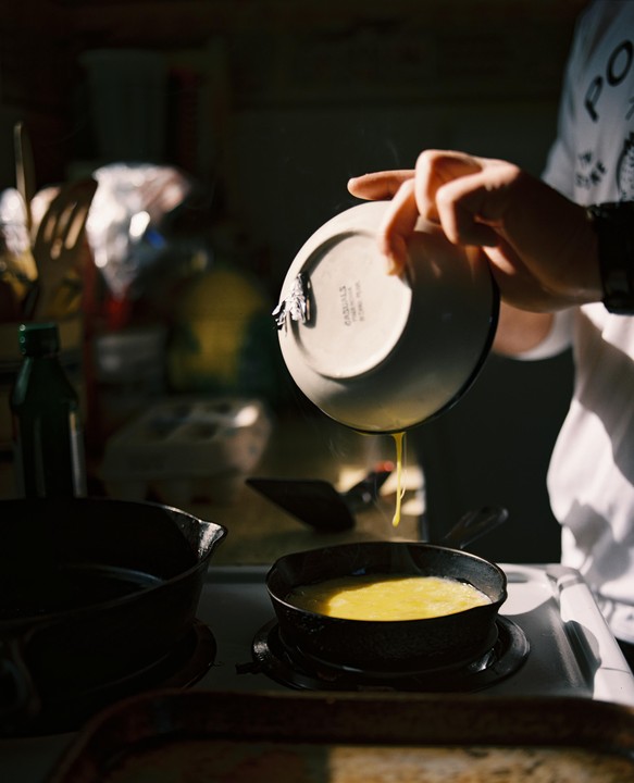 A person pours scrambled eggs from a bowl into a hot pan on a stove.