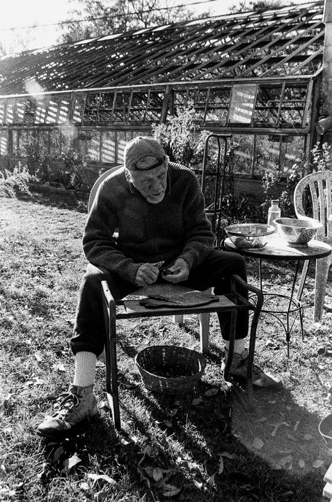 A man wearing a cap and sweater sits on a chair in a garden, peeling vegetables with a knife.