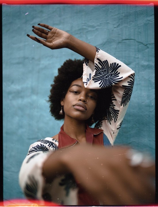 A woman with an afro and a nose ring poses with one arm raised and the other extended towards the viewer.