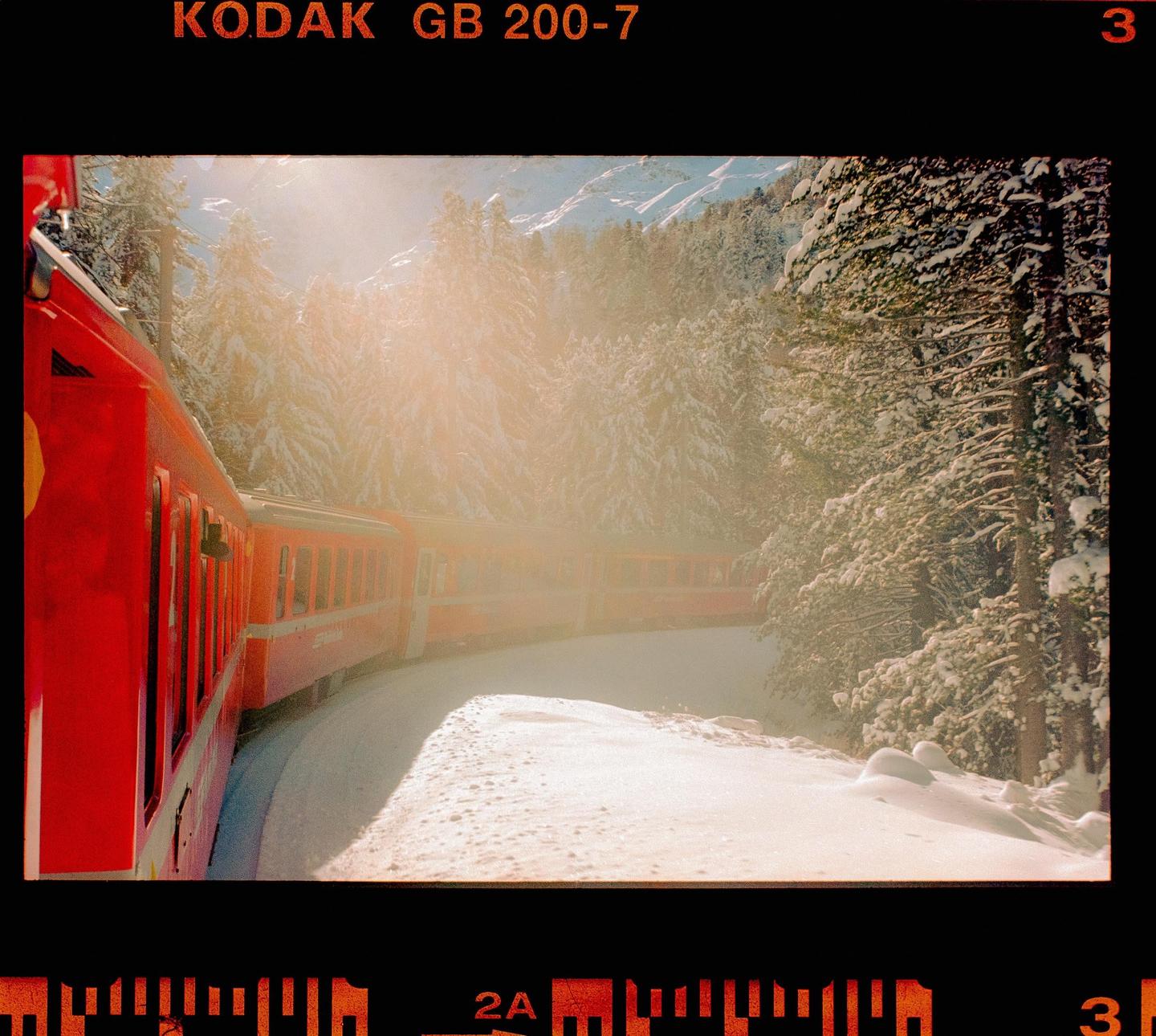 A red train travels through a snowy forest with sunlight shining through the trees.