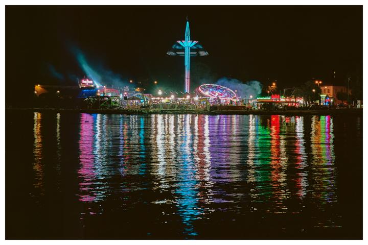 A fairground with rides and colorful lights is reflected in the water at night.