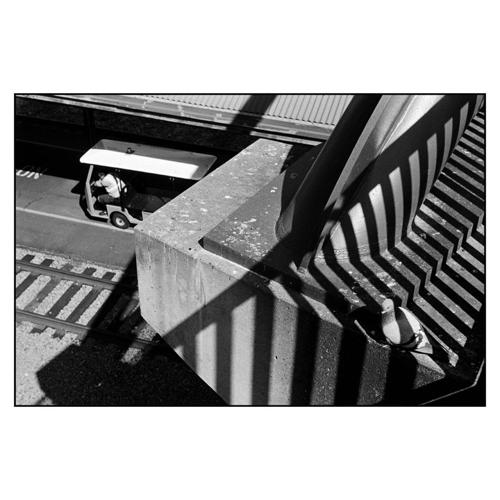 A man drives a golf cart past train tracks and a concrete structure with strong shadows.