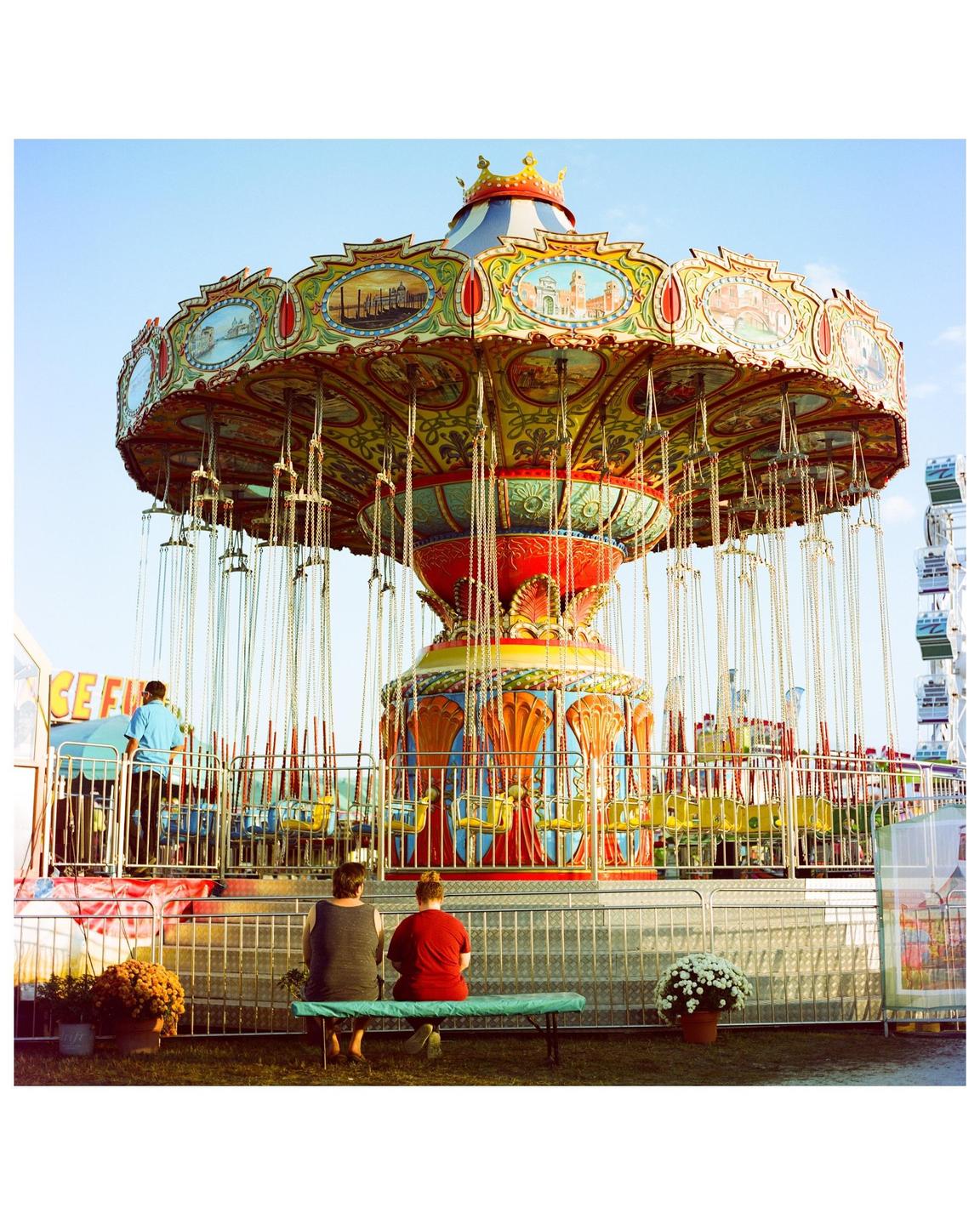 Two people sit on a bench in front of a colorful swing ride at a fair.