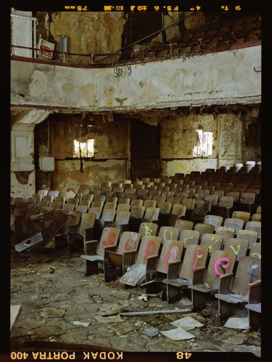 An abandoned auditorium with rows of wooden seats and a balcony is illuminated by light from windows.